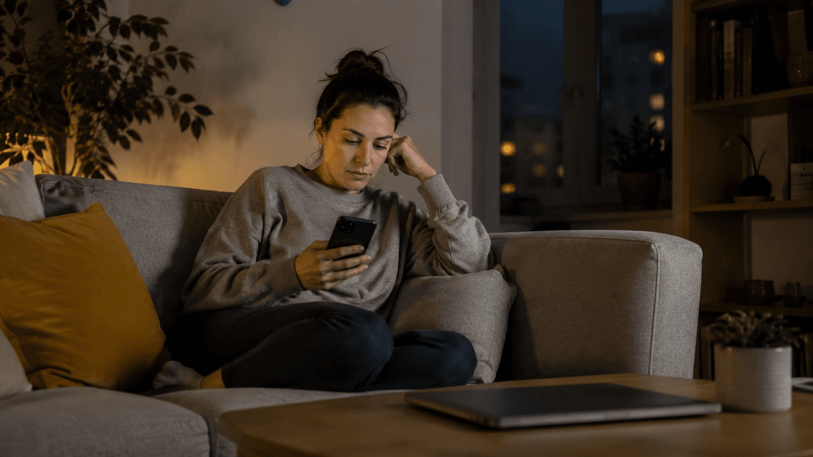 A person sitting on a couch, phone in hand, laptop closed on the table nearby, evening light