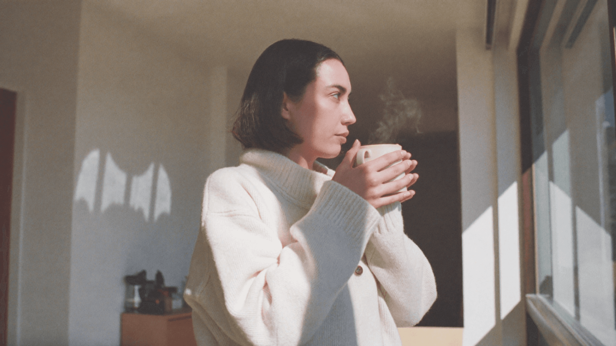 Person sitting by a window holding a mug, looking outside in morning light