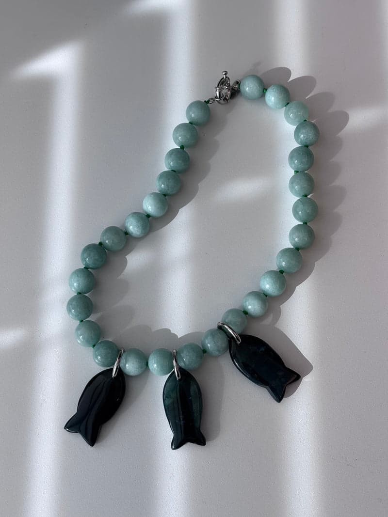 A pale green bead necklace with three dark green carved fish pendants, laid on a white surface in striped light