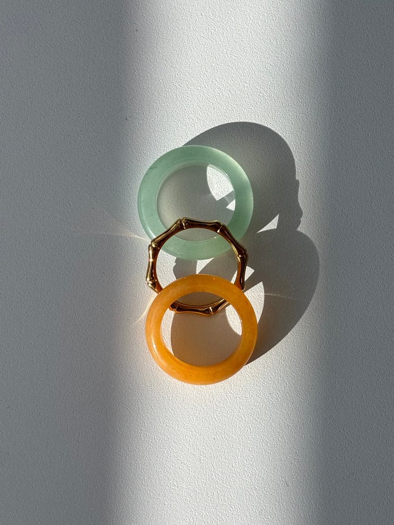 Two stone ring bands, one pale green and one orange, and a thin gold bamboo-shaped ring laid in a line in bright striped sunlight