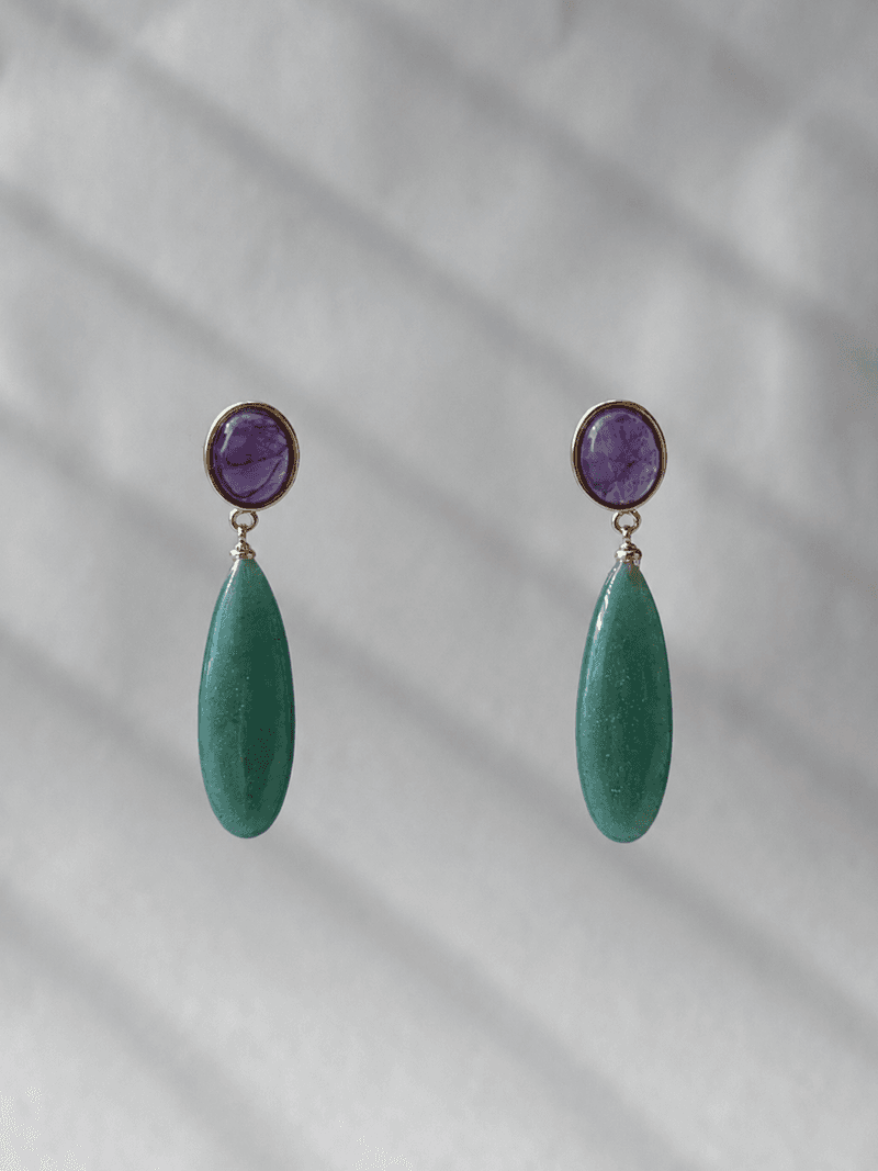 A pair of drop earrings, each a round purple stone in a gold bezel above a long green teardrop, on a white surface in striped light