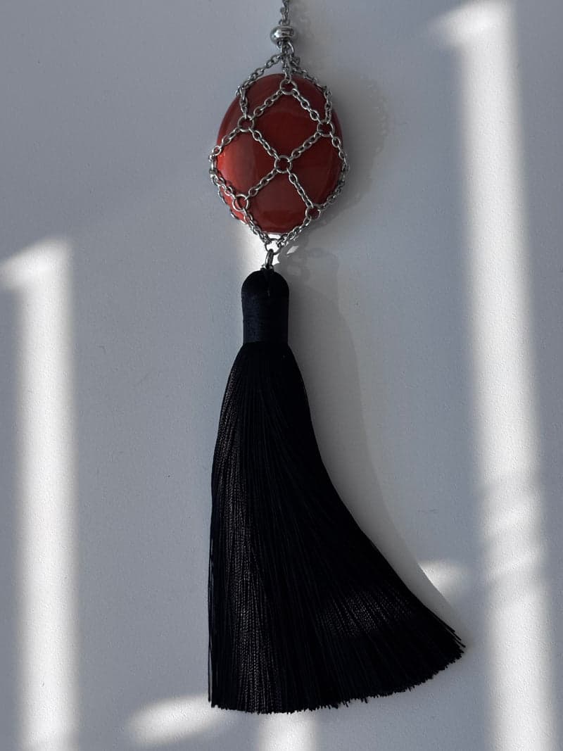 A brick red oval stone held in a silver chain mesh above a long black tassel, against a white wall in striped light