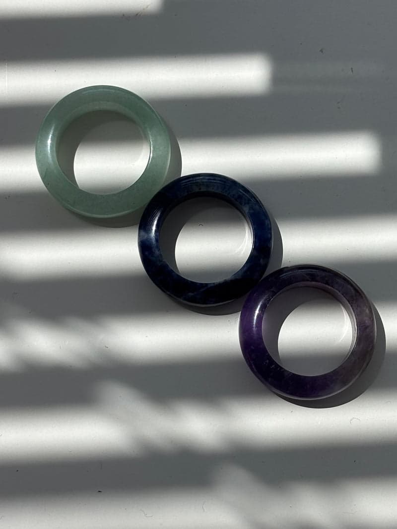 Three stone band rings laid in a diagonal line in striped sunlight: pale green, dark blue and purple