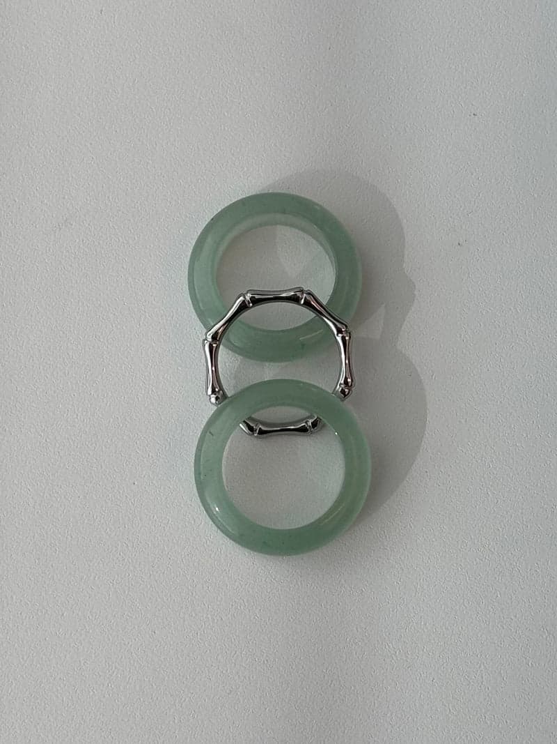 Two pale green stone rings and a thin silver-toned bamboo-shaped ring laid in a vertical line on a white surface
