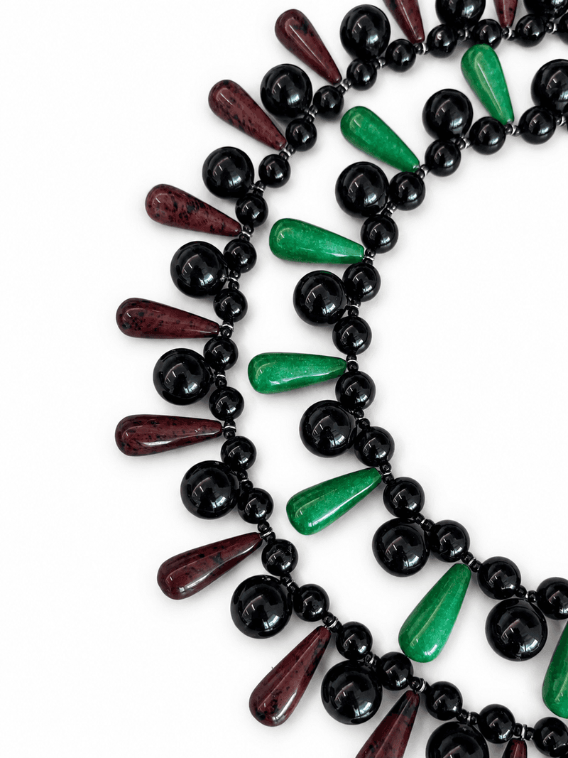 Two bib necklaces overlapping, one with green teardrop beads and one with dark red teardrop beads, both with round black beads