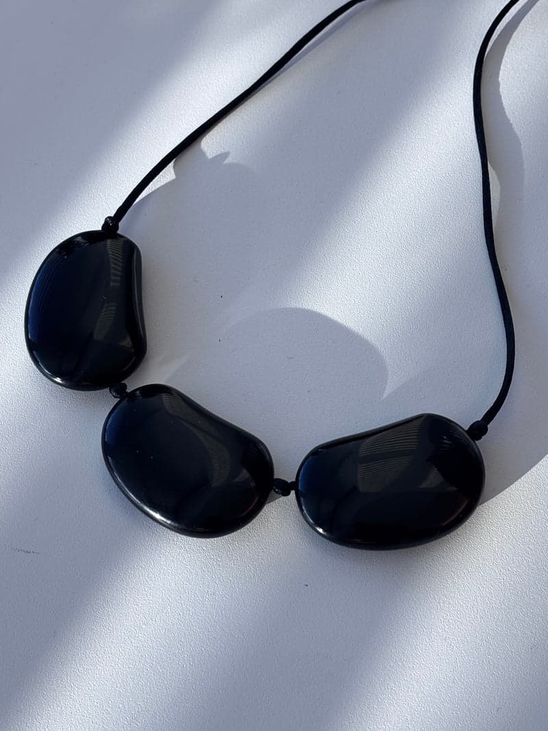 Three large flat glossy black oval stones strung on a black cord, lying on a white surface in striped light