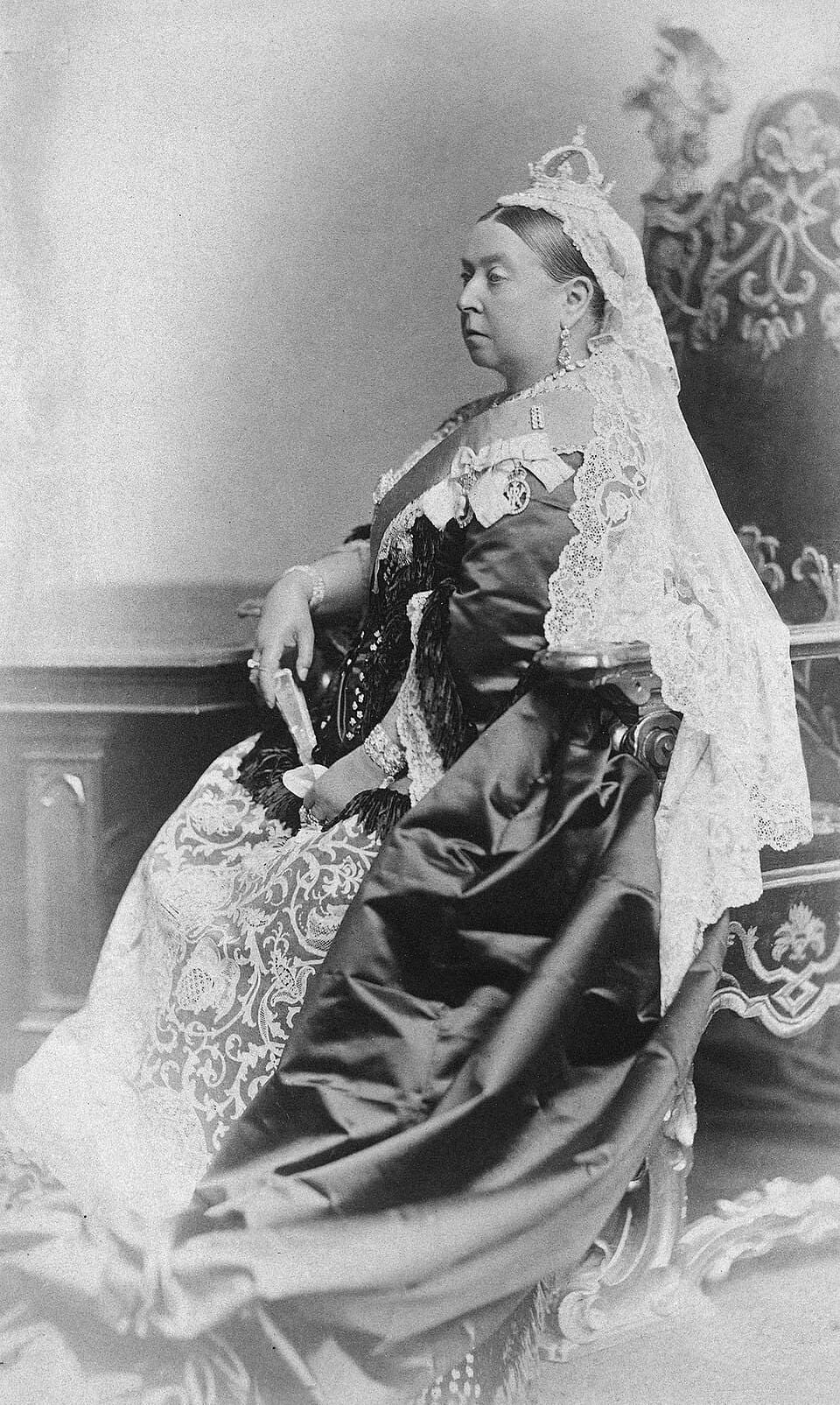 A black and white photograph of Queen Victoria seated, wearing a small crown and a long white lace veil