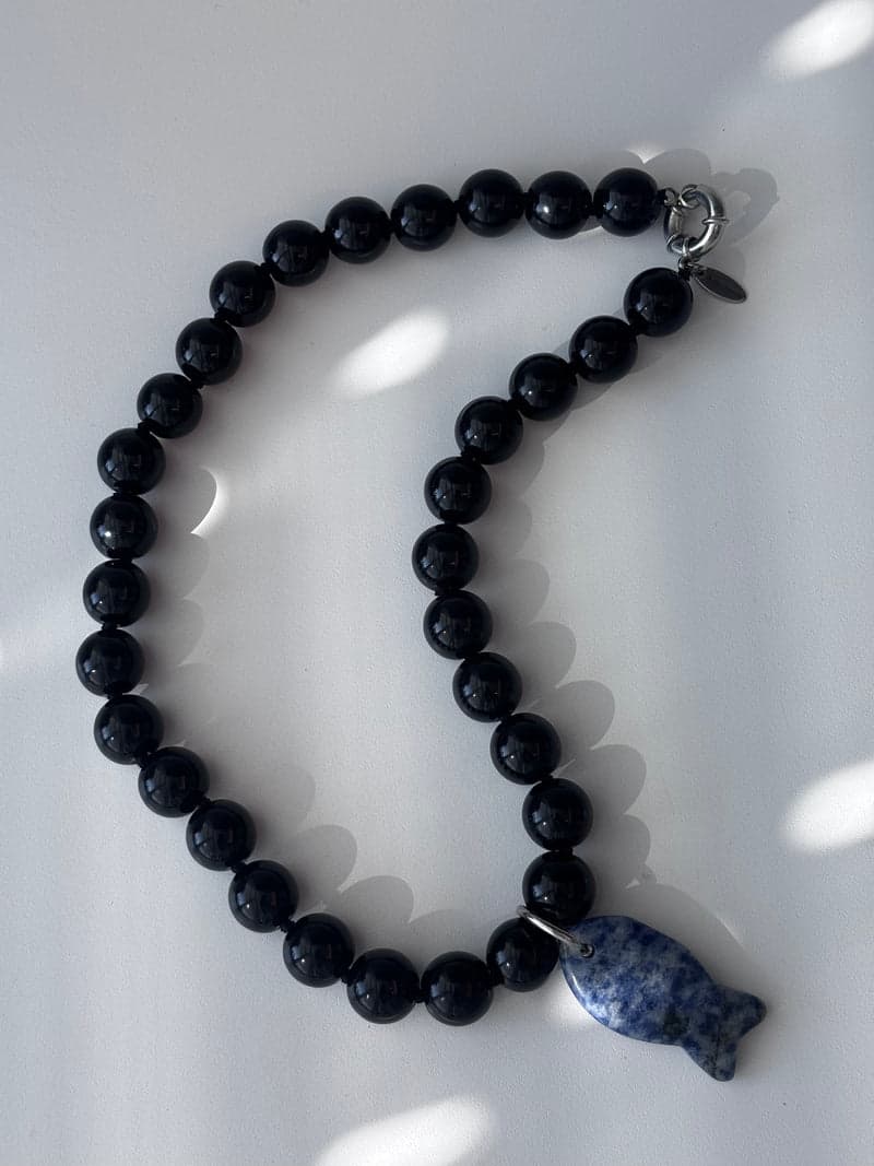 A strand of large glossy black beads in a curve on a white surface, with a blue and white mottled carved fish pendant at the bottom