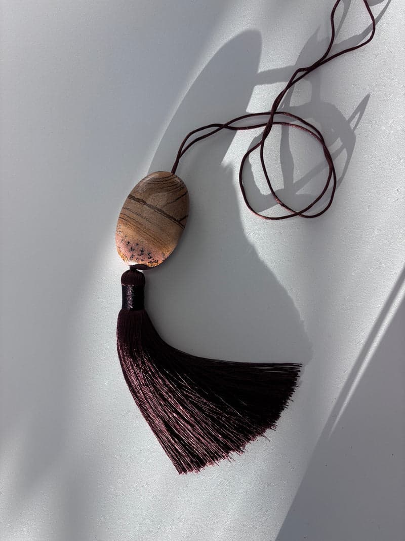 A flat oval stone in tan, grey and brown with fine dark branching marks, hanging from a brown cord above a deep brown tassel