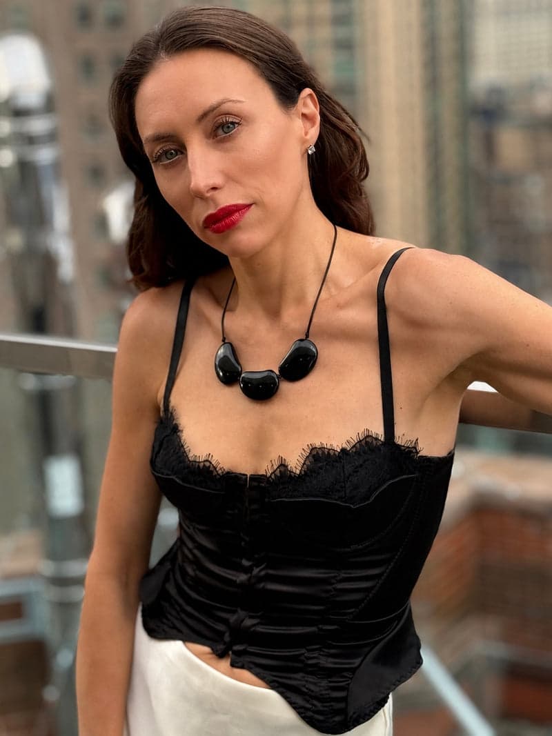 A woman on a city balcony in a black lace top wearing a necklace of three large dark oval stones on a black cord