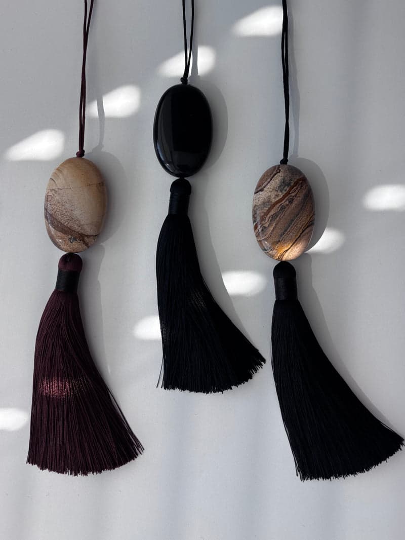 Three tasselled pendants side by side: a cream patterned stone, a black glassy stone, and a brown patterned stone