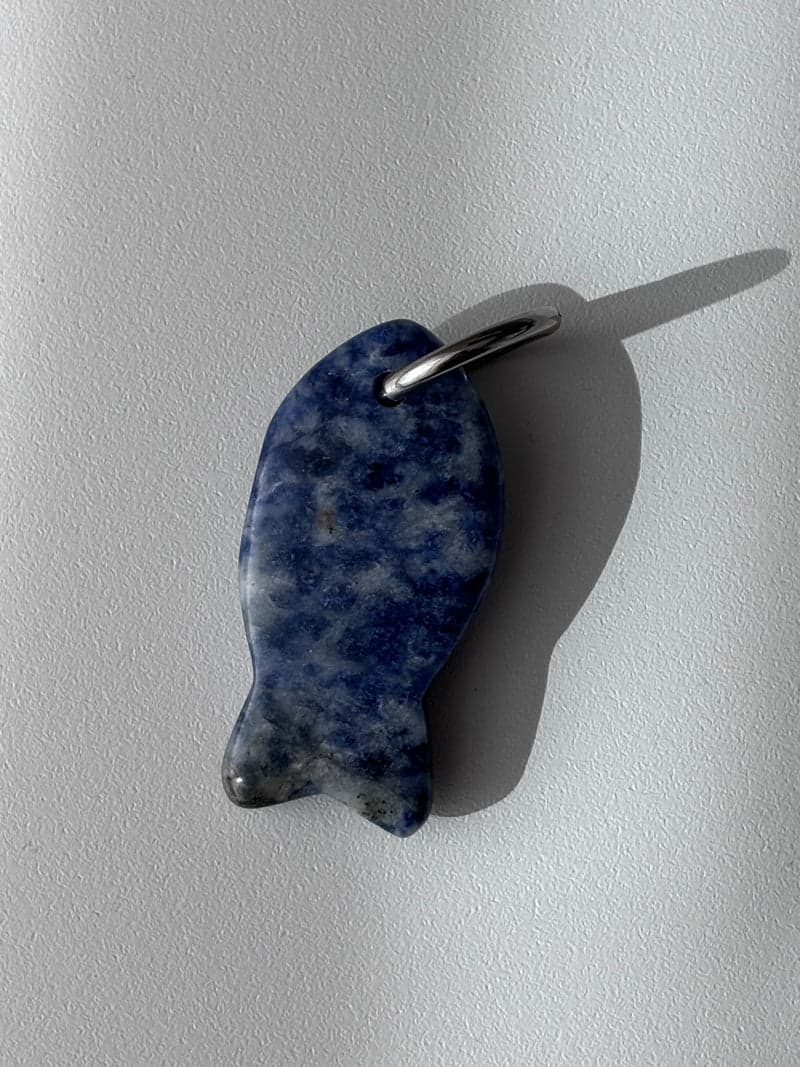 A small carved fish pendant in mottled blue and white stone with a silver ring, on a white surface in window light