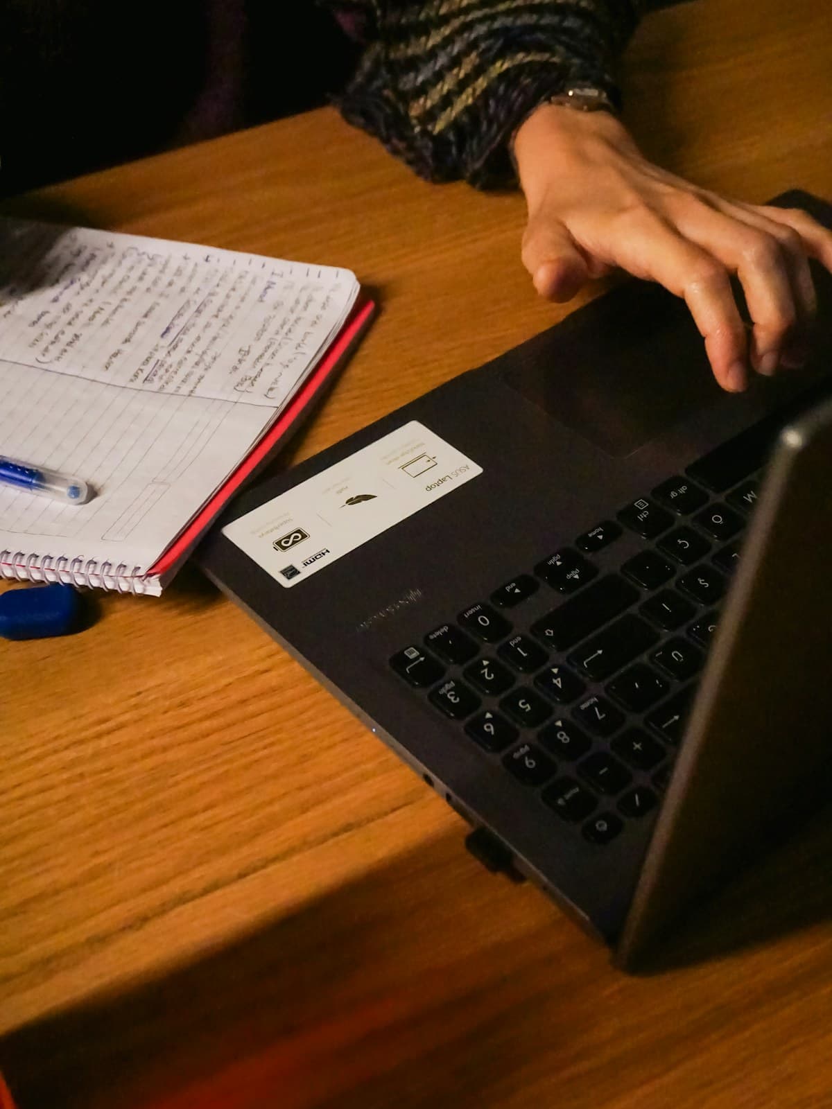 Manos escribiendo en un portátil sobre un escritorio de madera junto a un cuaderno abierto