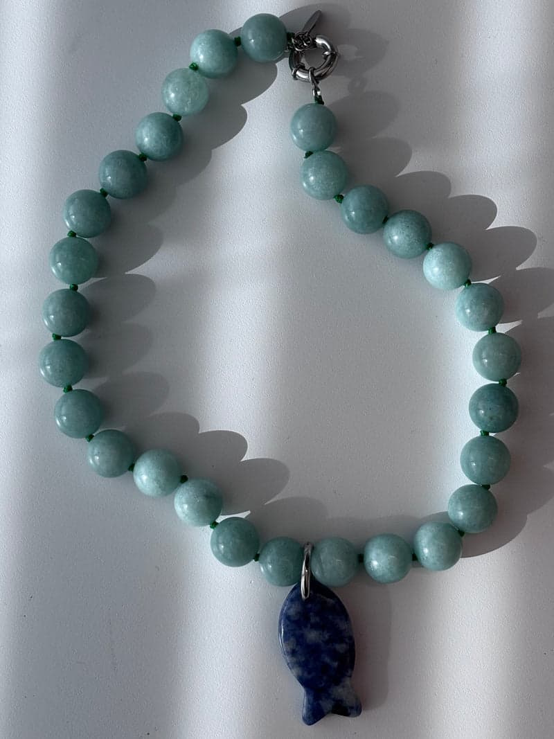 A pale green bead necklace laid on a white surface with a single blue and white carved fish pendant at the bottom