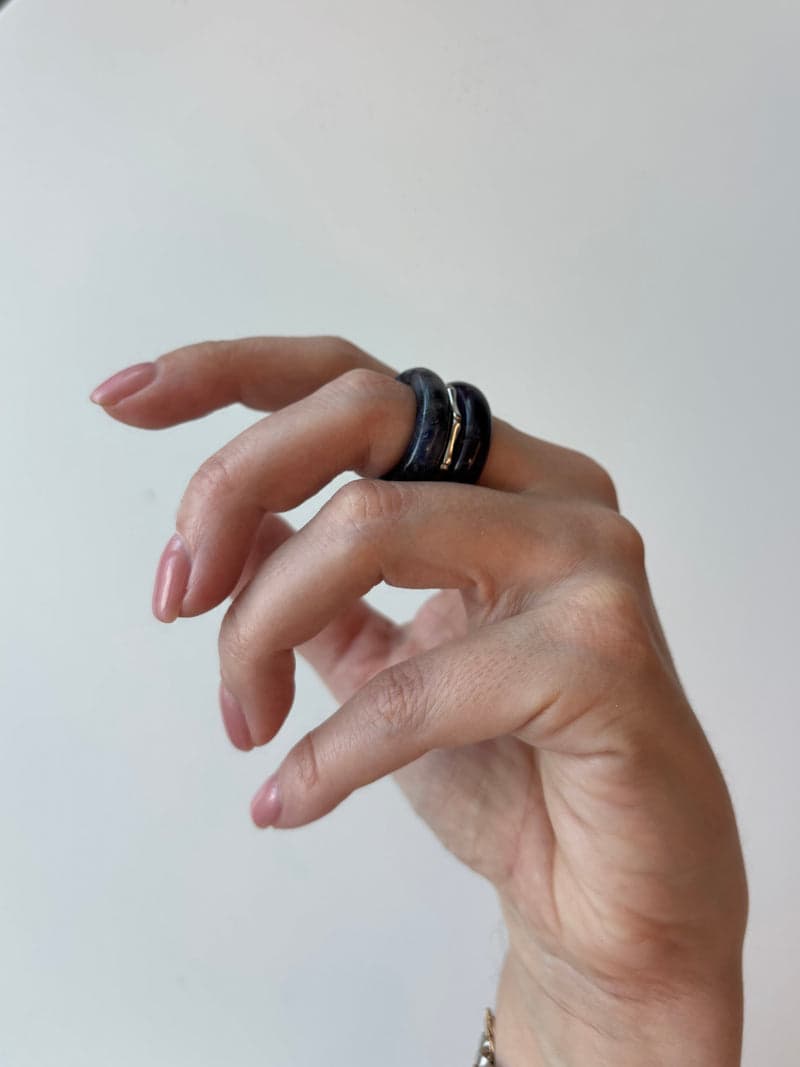 A raised hand against a pale background wearing the dark blue and purple stone rings stacked on one finger