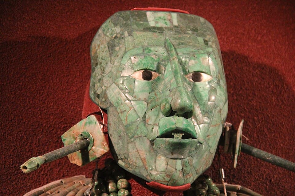 A green mosaic mask made of many pieces of jade, with white shell eyes and dark pupils, against a red background