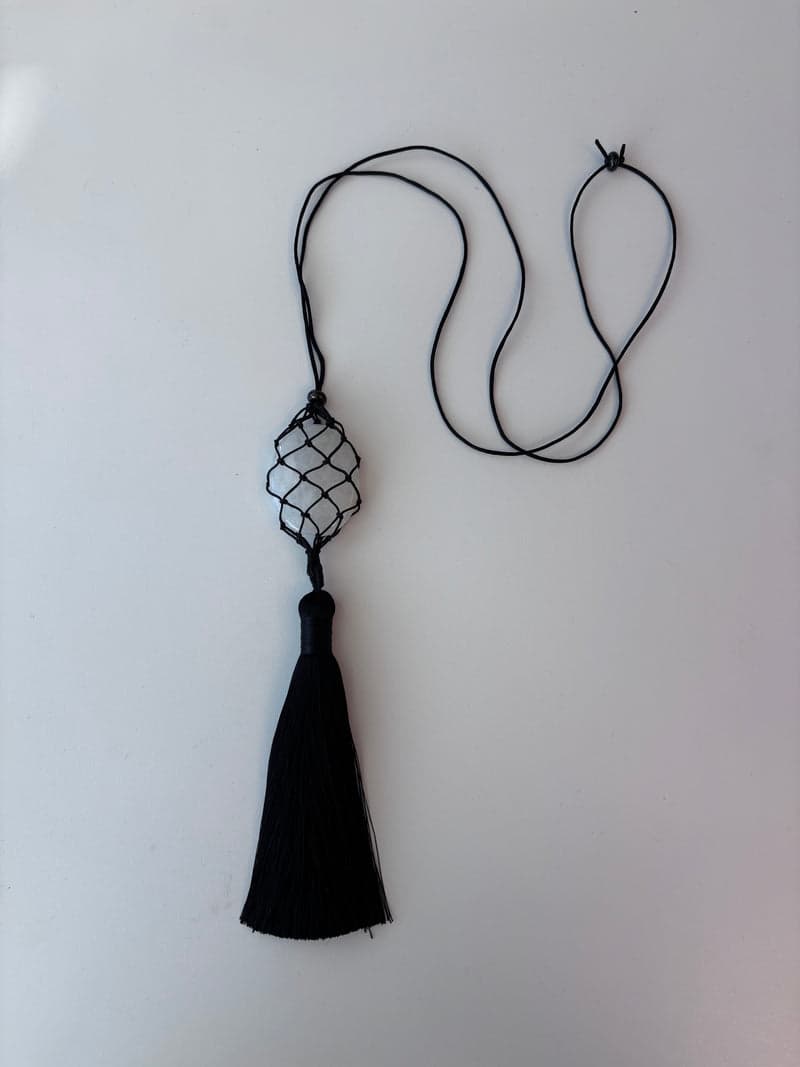 A white quartz stone held in a black knotted cord net on a long black cord, above a long black tassel, on a white surface