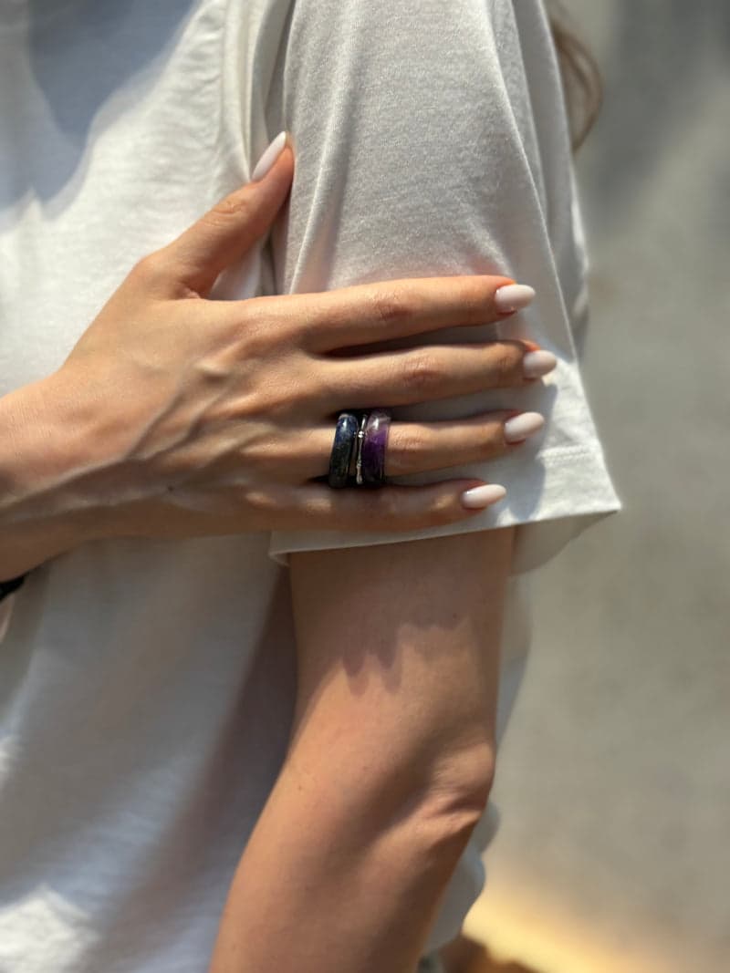 A hand resting on a bare upper arm against a white t-shirt, wearing the purple and dark blue stone rings stacked on one finger