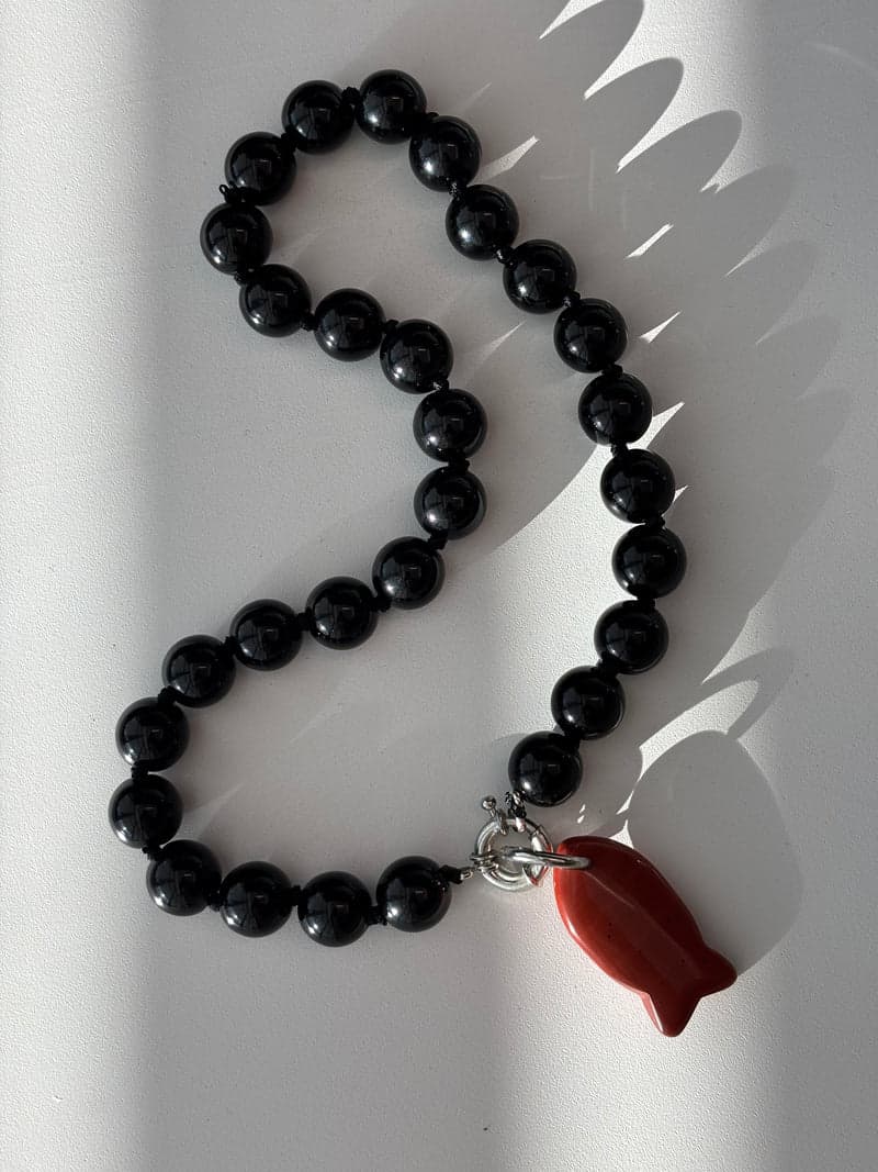 A strand of large glossy black beads laid in a curve on a white surface, with a small red carved fish pendant at the bottom