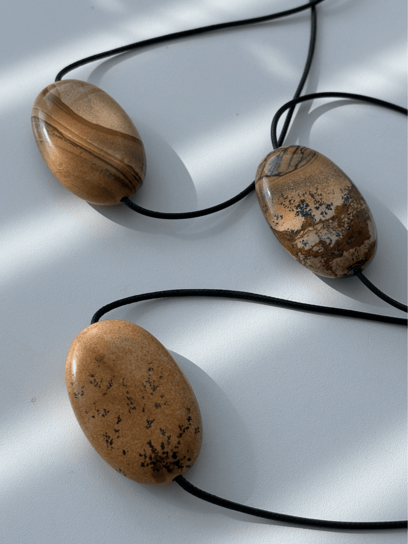 Three large flat oval pendants in tan and brown stone with dark branching marks, on black cords on a white surface