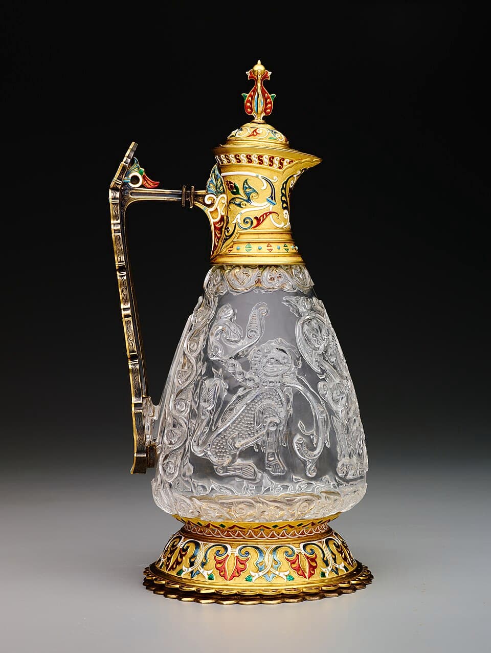 A clear carved rock crystal jug with an ornate enamelled gold lid, handle and foot, on a dark background