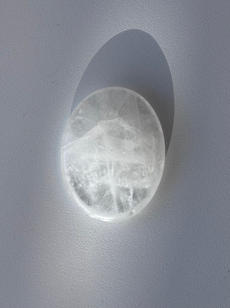 A polished oval of clear quartz with white internal veils, lying on a white surface and casting a soft shadow