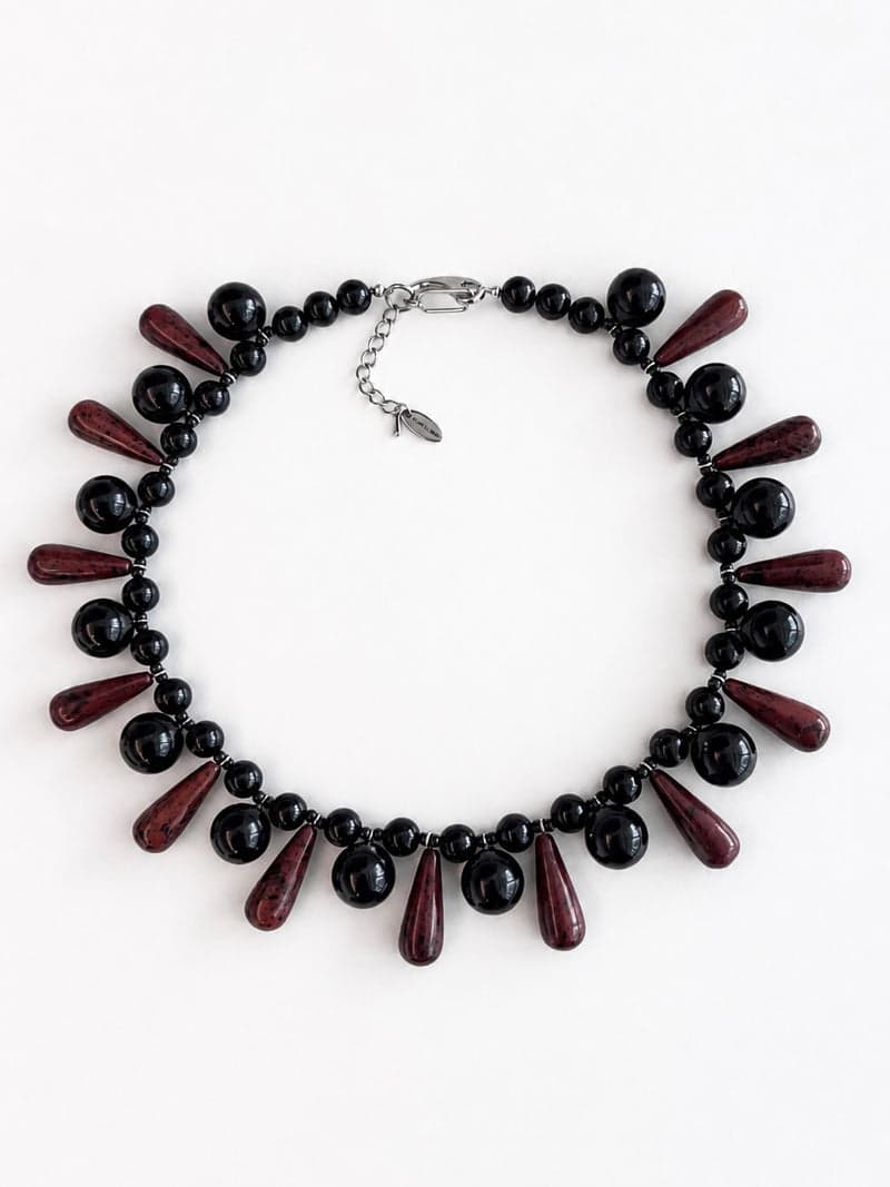 A bib necklace of round glossy black beads alternating with long dark red teardrop beads, on a white background