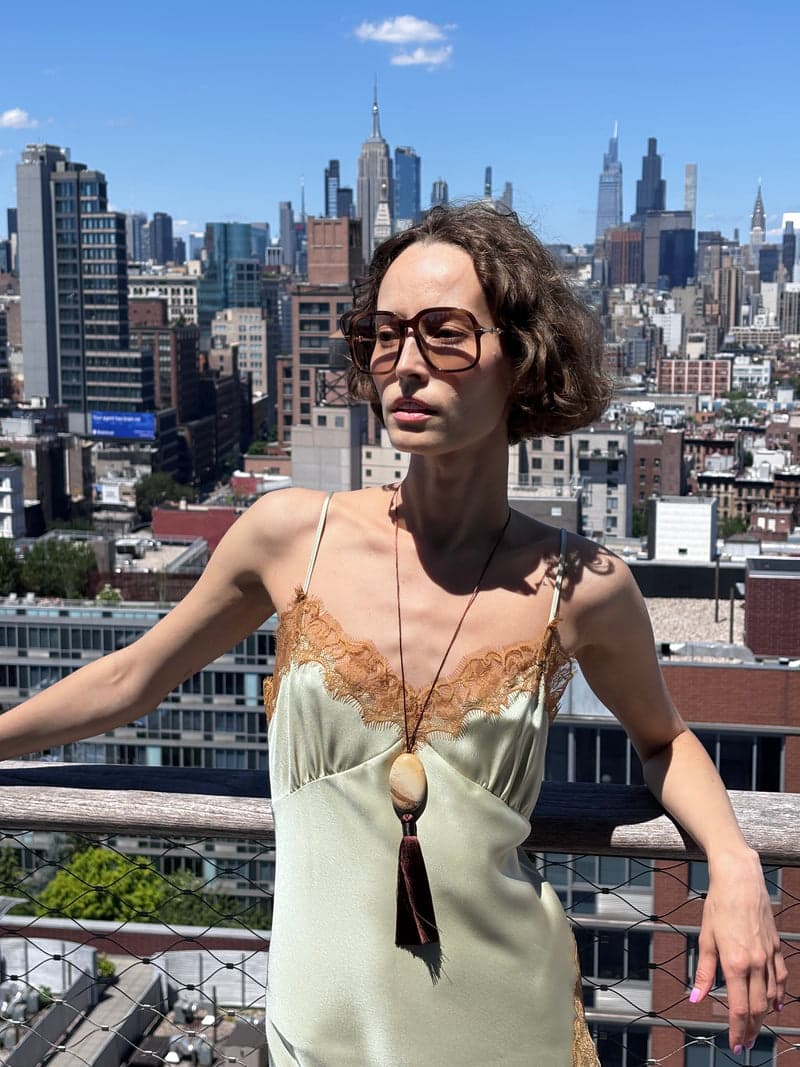 A woman on a city rooftop in a pale green slip dress wearing a long cord with a tan oval stone and a brown tassel