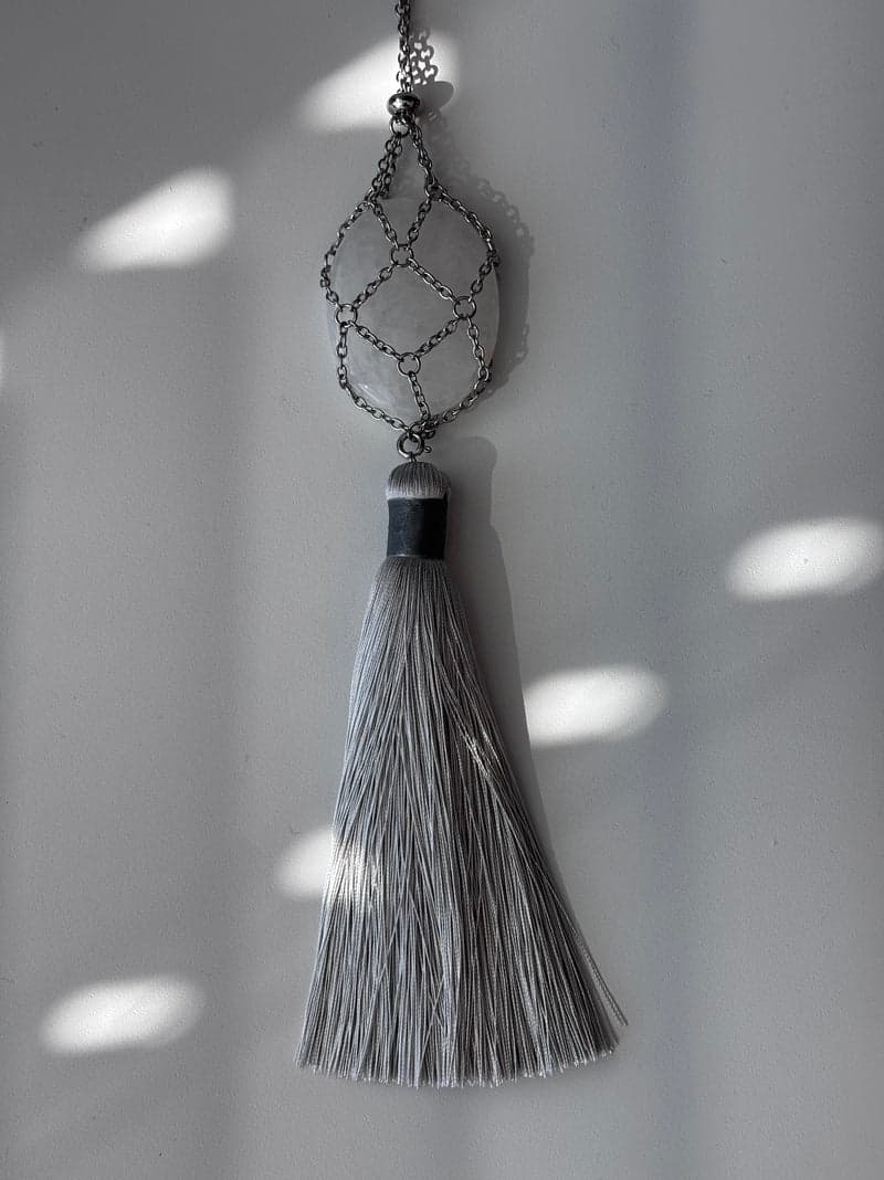 A pale, clouded quartz oval held in a silver chain mesh above a long grey tassel, against a white wall in striped light