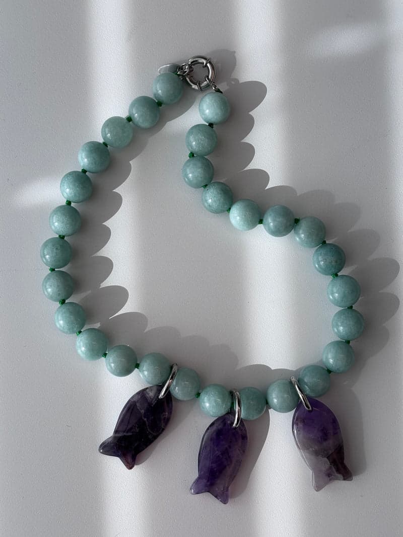 A pale green bead necklace with three purple carved fish pendants, laid on a white surface in striped light