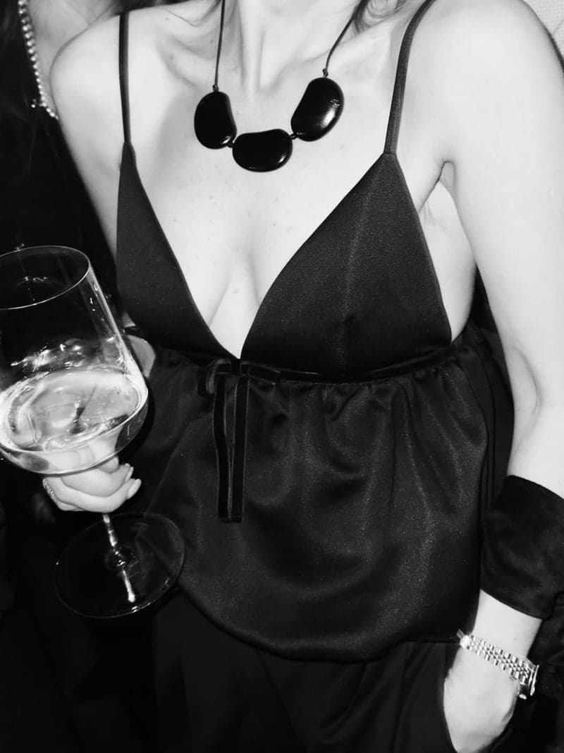 A black and white photograph of a woman in a black slip top holding a wine glass, wearing a necklace of three large dark oval stones