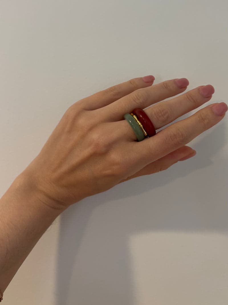 A hand held against a pale wall wearing the red and pale green stone rings stacked on one finger