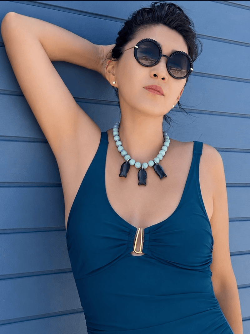 A woman in sunglasses and a teal swimsuit against a blue wall, wearing a pale green bead necklace with three dark fish pendants