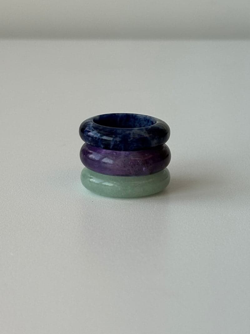 Three stone band rings stacked on a white surface: dark blue on top, purple in the middle, pale green below
