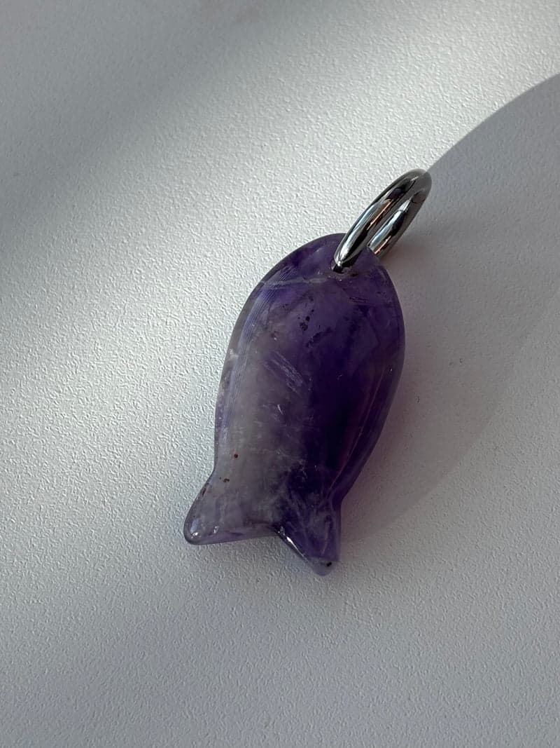 A small carved fish pendant in translucent purple stone with a silver ring, on a white surface in window light
