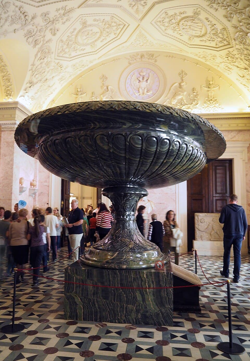 An enormous shallow green stone bowl on a pedestal in a marble hall of the Hermitage, with visitors around it