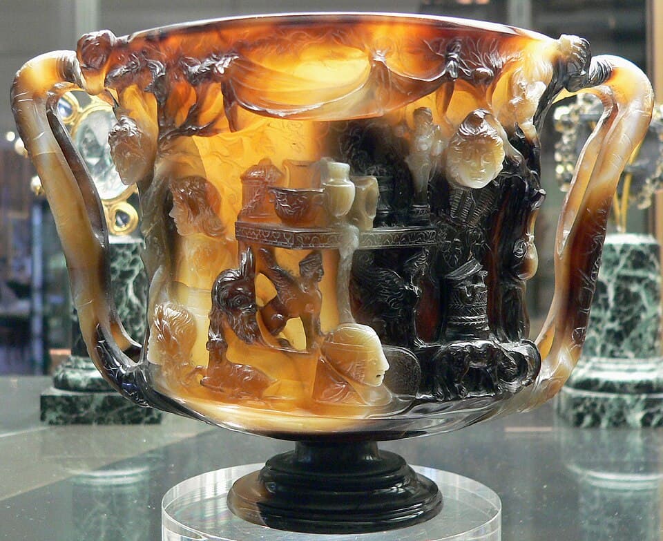 An ancient two-handled cup carved from banded brown, cream and white stone, with relief scenes, in a museum case