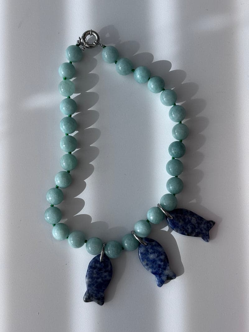 A pale green bead necklace with three blue and white mottled carved fish pendants, laid on a white surface in striped light