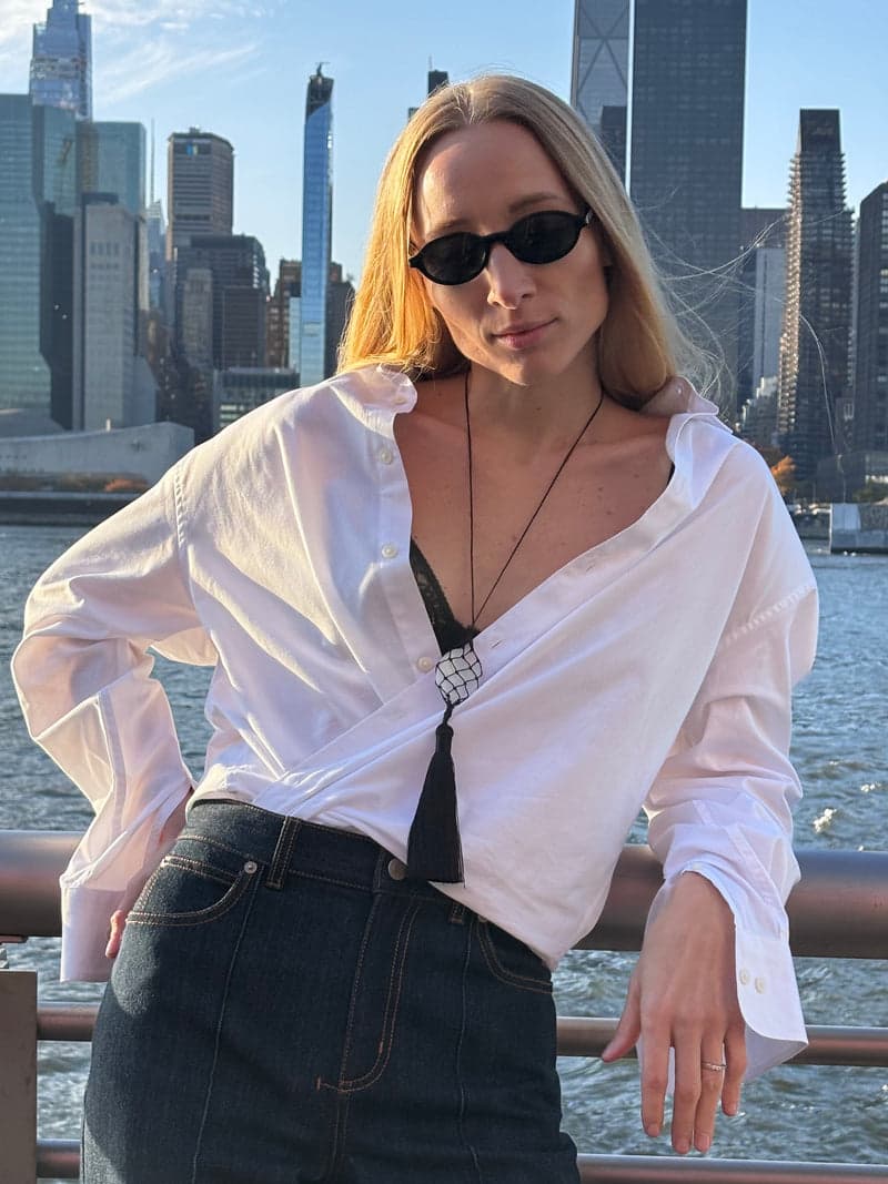 A woman in sunglasses and an open white shirt by a city waterfront, wearing the long cord necklace with the white quartz stone and black tassel