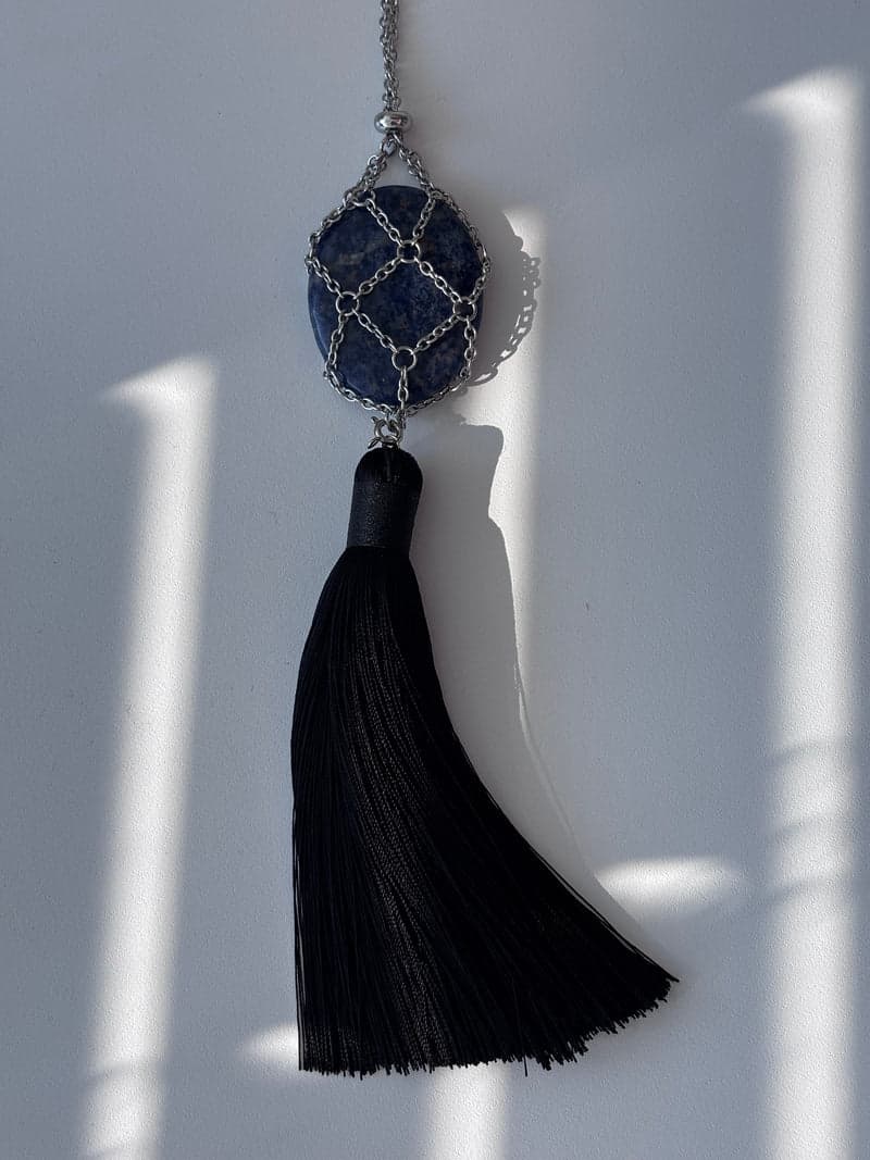 A mottled blue and white oval stone held in a silver chain mesh above a long black tassel, against a white wall in striped light