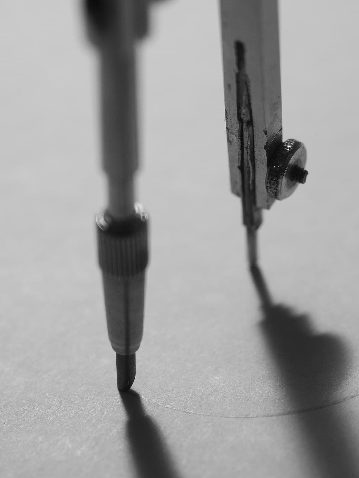 Un compás metálico de dibujo trazando un círculo sobre papel blanco, en blanco y negro