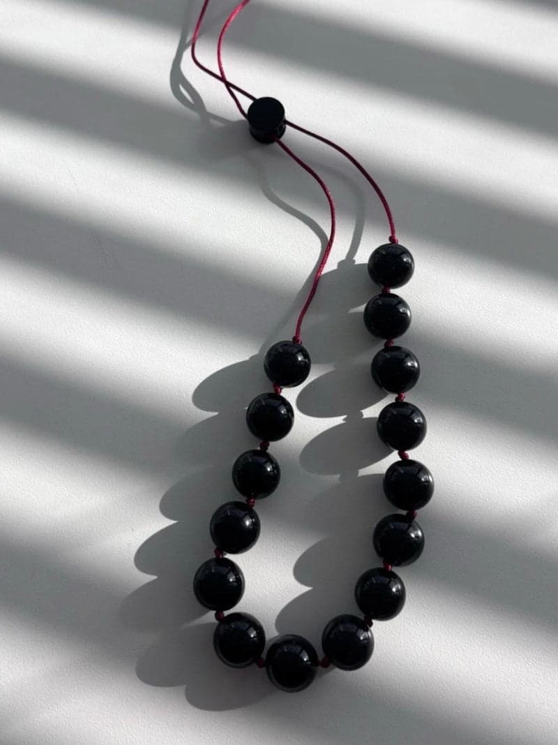 A necklace of large glossy black round beads strung on a thin red cord, laid in a loop on a white surface in striped window light