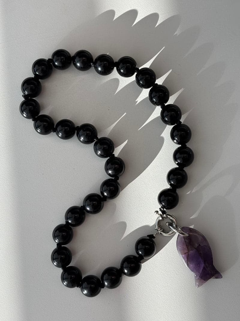 A strand of large glossy black beads in a curve on a white surface, with the purple carved fish pendant at the bottom