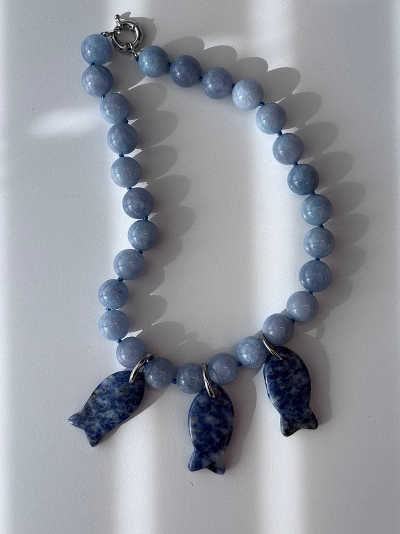 A pale blue bead necklace with three blue and white mottled carved fish pendants, laid on a white surface in striped light