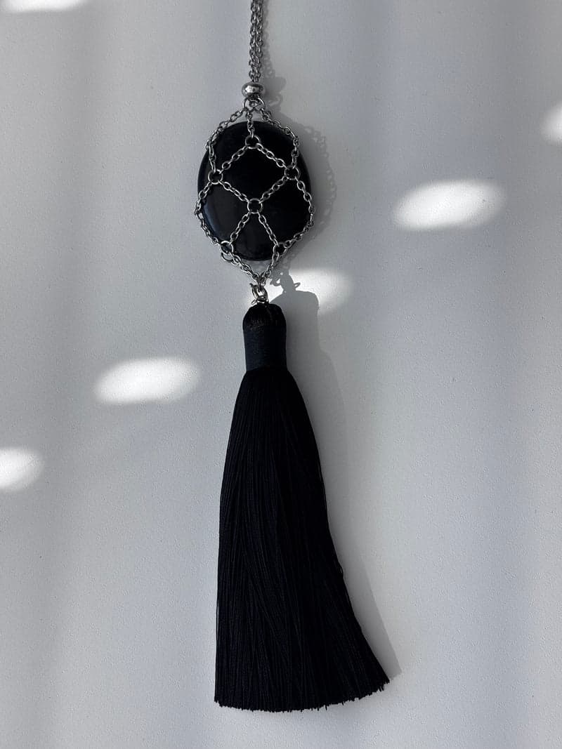 A black obsidian oval held in a silver chain-mesh net above a black tassel, hanging against a white wall