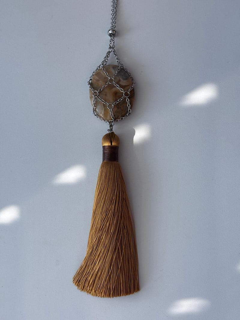 An oval banded agate in cream, tan and grey held in a silver chain mesh above a long golden brown tassel